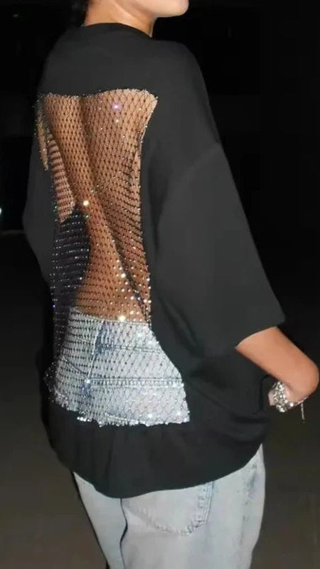 A person shown from the back wearing an oversized black top with a large back panel made of sheer mesh and sequins in warm beige/tan and silver tones, paired with light blue jeans.